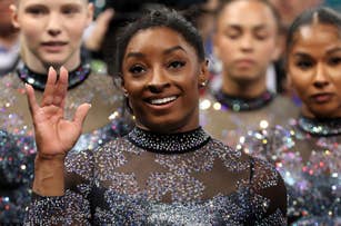 Simone Biles waves while standing with her teammates. They are dressed in sequined gymnastics leotards during a competition