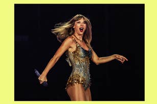 Taylor Swift performing on stage in a shimmering, sleeveless dress. She holds a microphone in her right hand and looks playfully shocked
