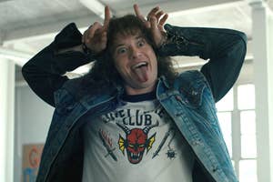 Joseph Quinn, as Eddie Munson, playfully posing with tongue out and hands making horns, wearing a denim jacket and a Hellfire Club t-shirt