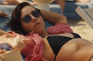 Aubrey Plaza is lounging on a sunbed at the beach, wearing sunglasses, a dark bikini, and an open shirt as Harper on The White Lotus